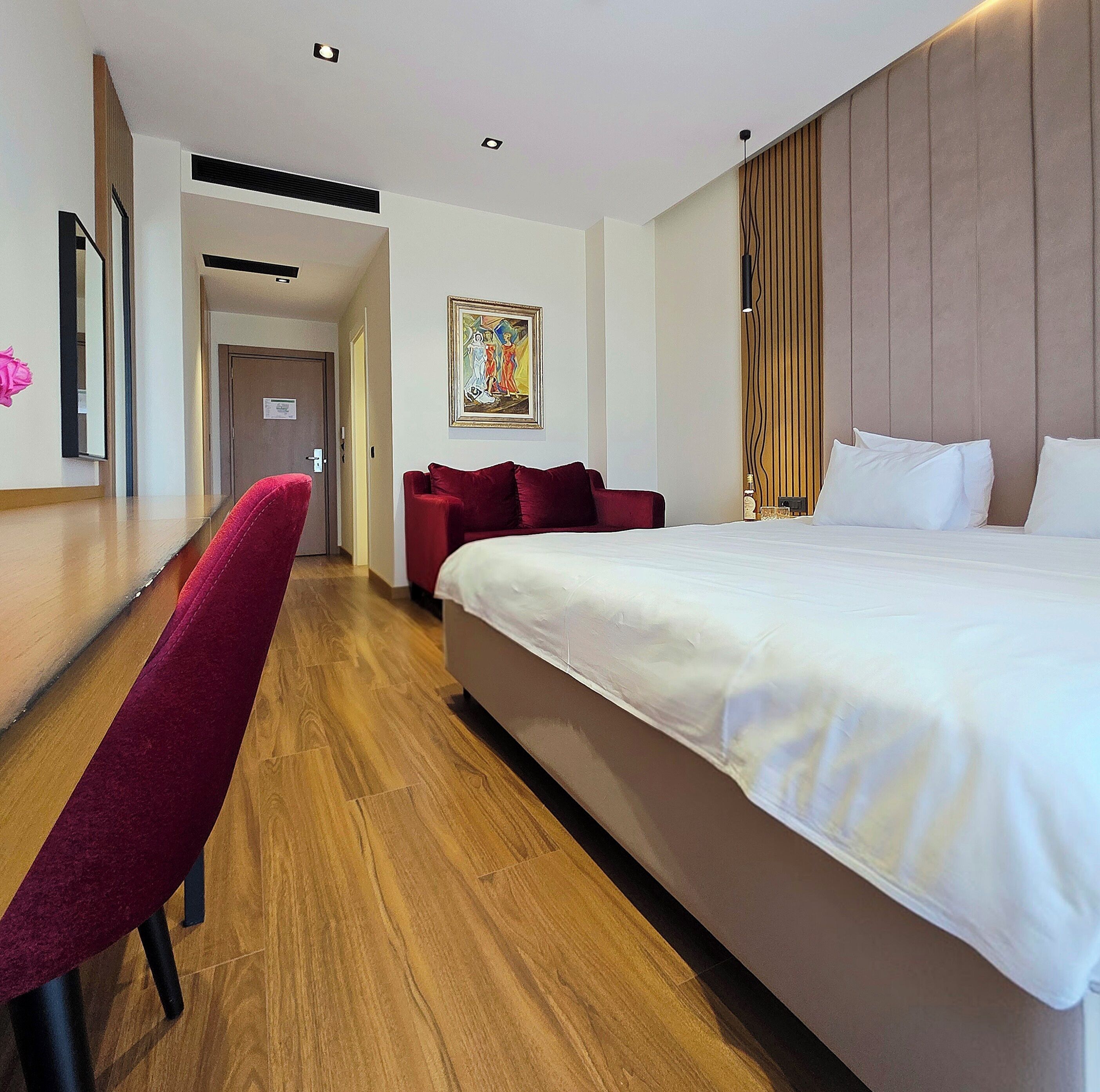 A hotel room with a large bed, a desk with a chair, a wooden floor, and a painting on the wall.
