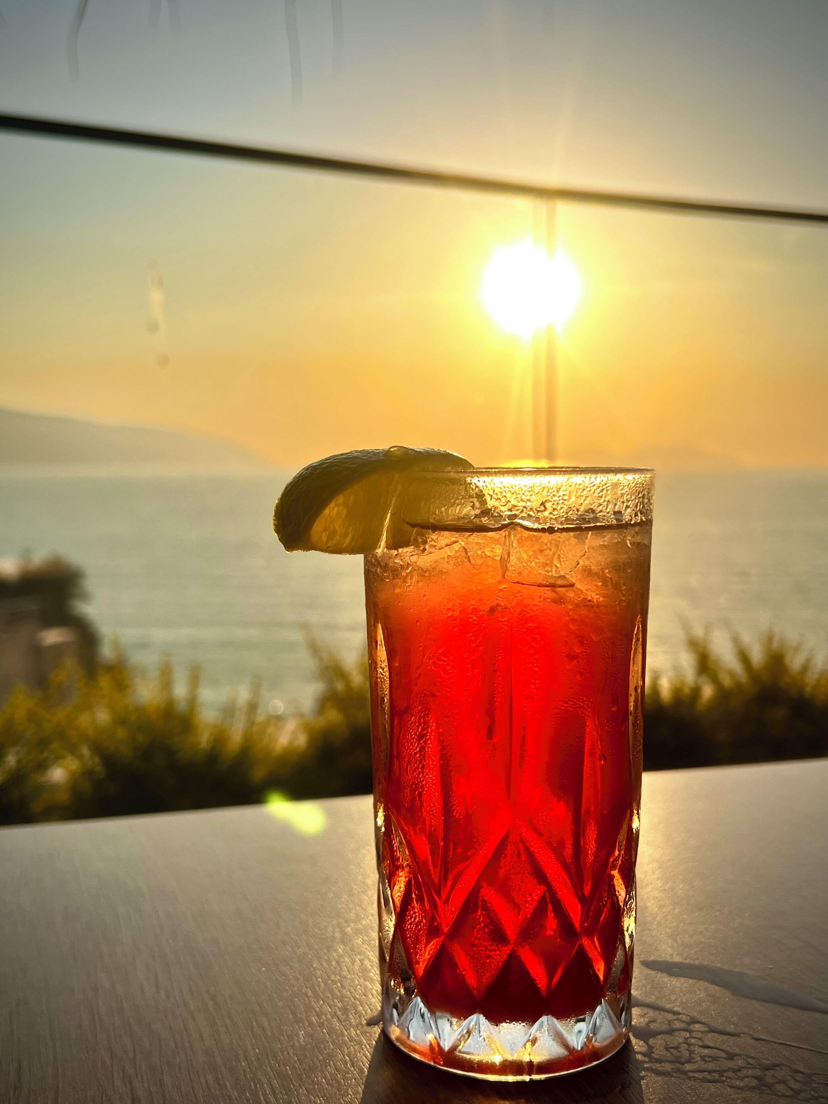 A glass of red beverage with a lemon slice on a table with a sunset view.