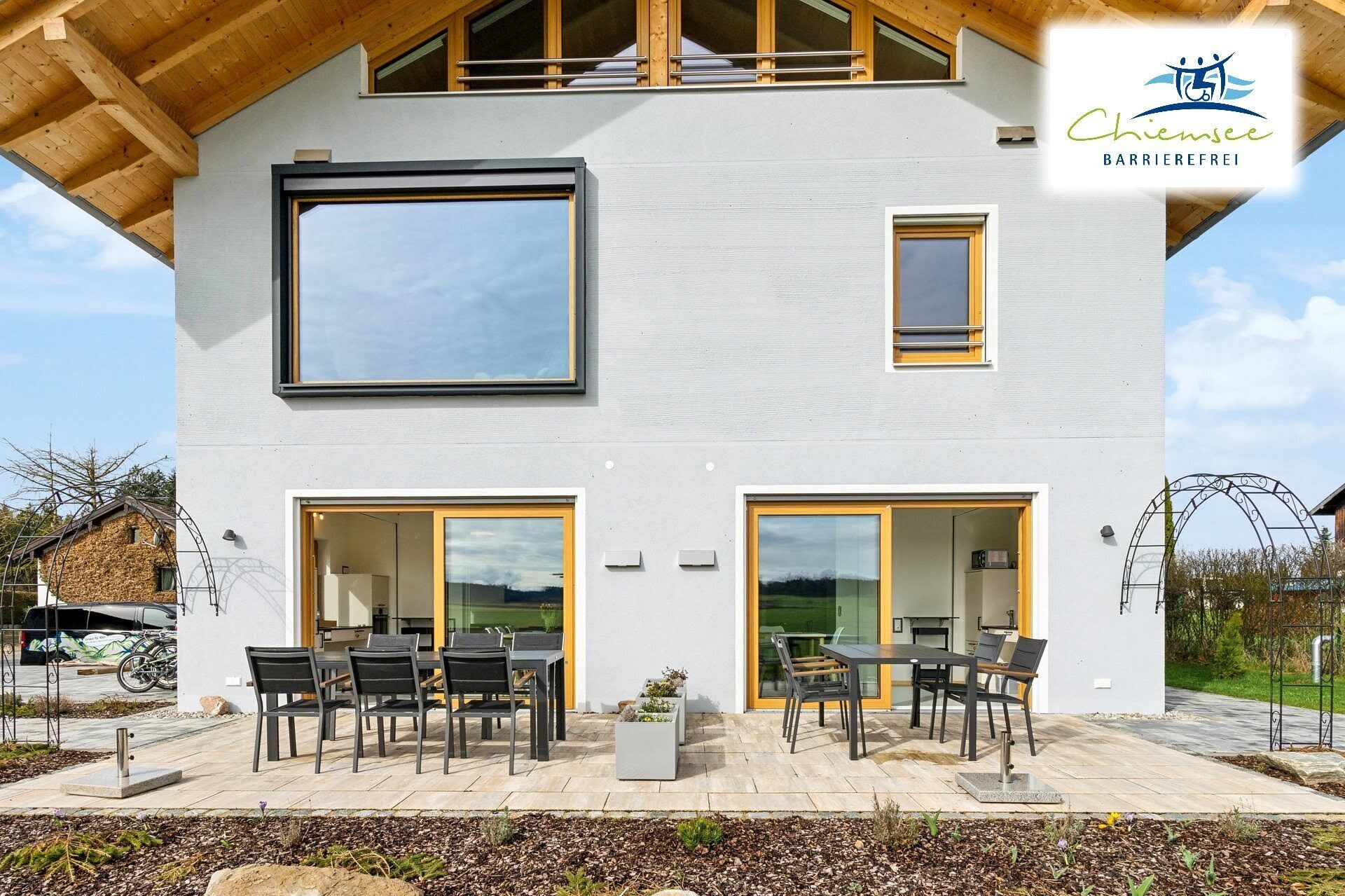 Une maison moderne à deux étages, dotée de grandes fenêtres, d’un balcon et d’un espace salon extérieur.