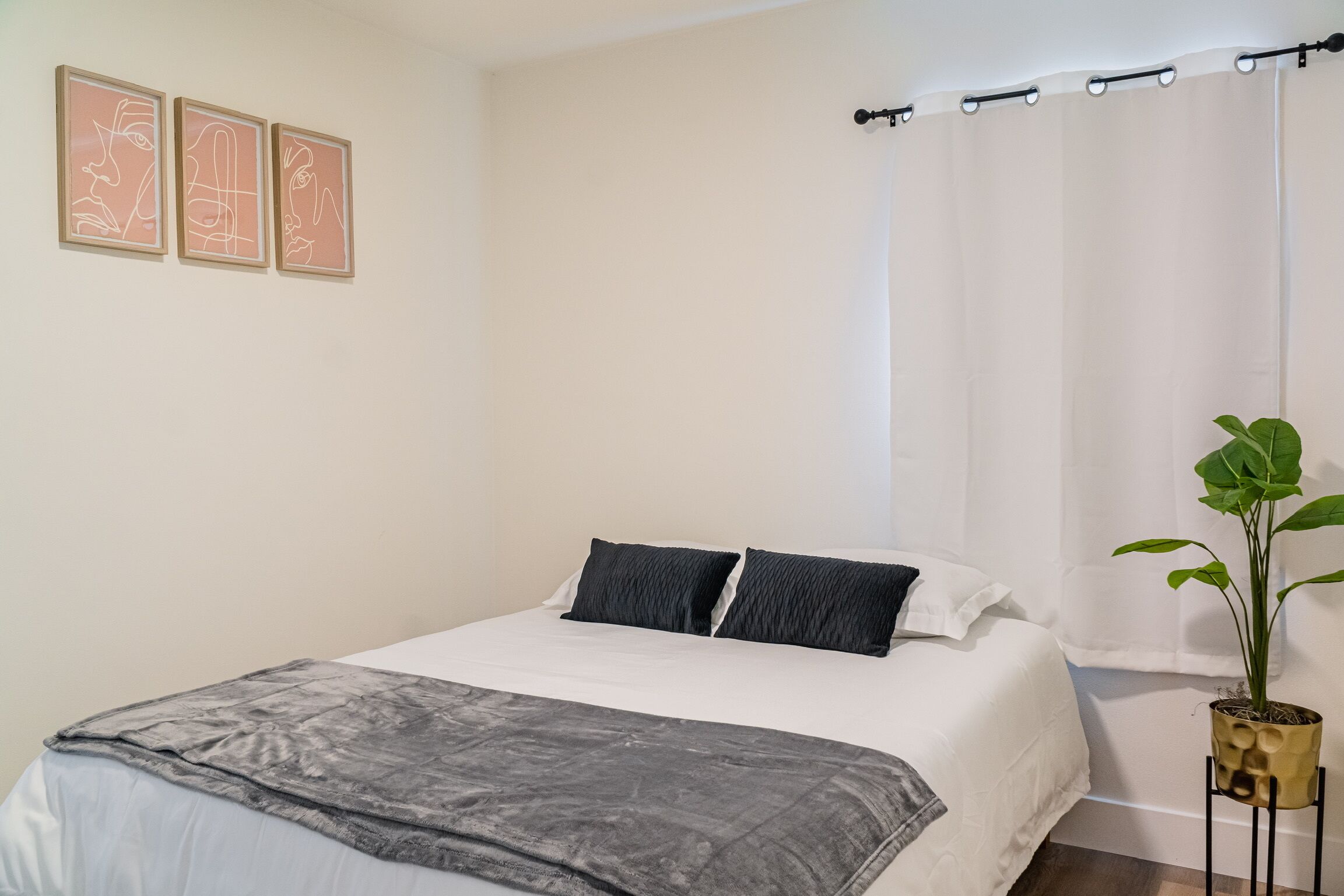 Una cama bien tendida con un edredón blanco, dos almohadas negras y una manta gris. En la pared hay tres cuadros enmarcados, una planta en maceta sobre un pedestal y una cortina blanca.
