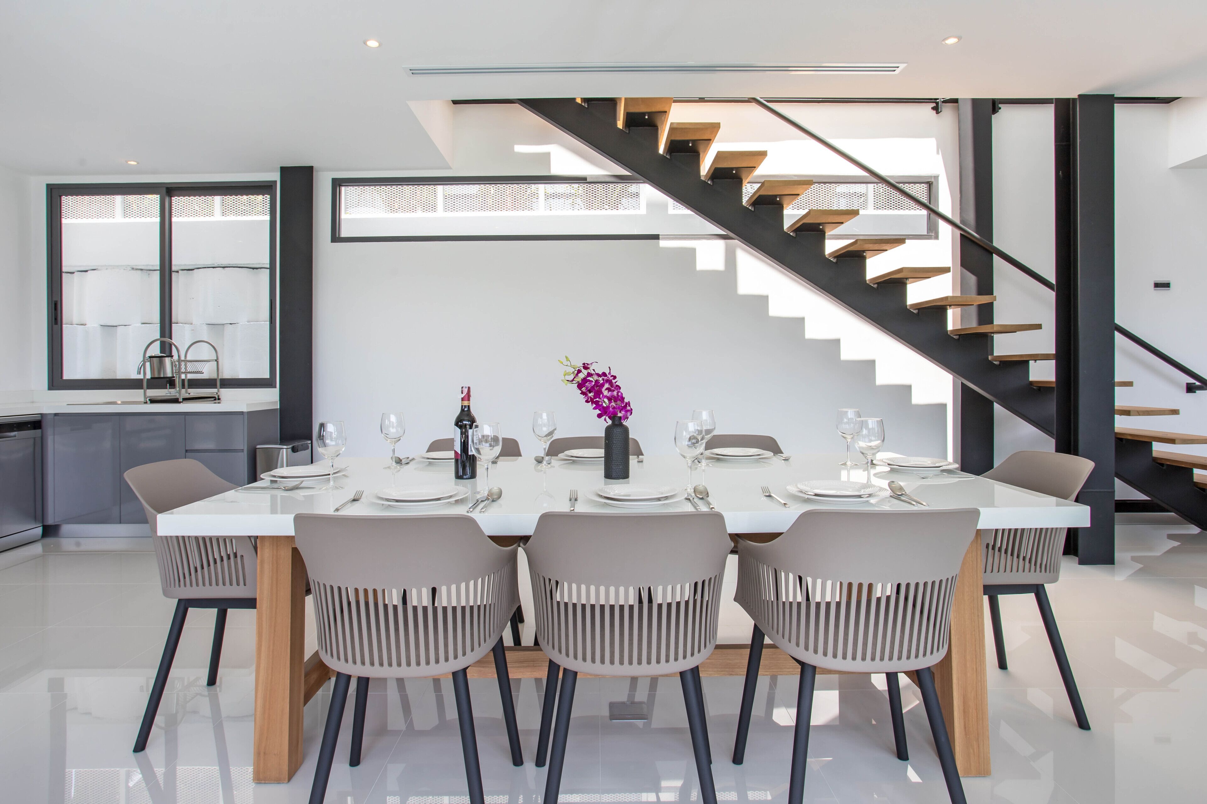 A modern dining area with a white table set for four, surrounded by grey chairs, and a staircase with wooden steps and black railings.