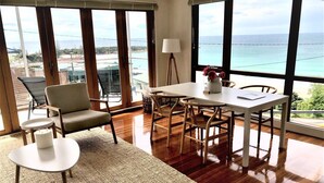 A spacious room with a large window offering a view of the ocean and a pool. There is a dining table with chairs, a sofa, and a lamp. - OUTSTANDING! WHITE HOUSE TASMANIA WINTER SPECIAL MIN STAY 2 NIGHTS TILL OCTOBER. (Boat Harbour Beach)