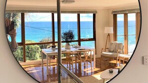 A spacious room with large windows offering a view of the sea, a round mirror on the wall, and a dining area with a table and chairs. - OUTSTANDING! WHITE HOUSE TASMANIA WINTER SPECIAL MIN STAY 2 NIGHTS TILL OCTOBER. (Boat Harbour Beach)