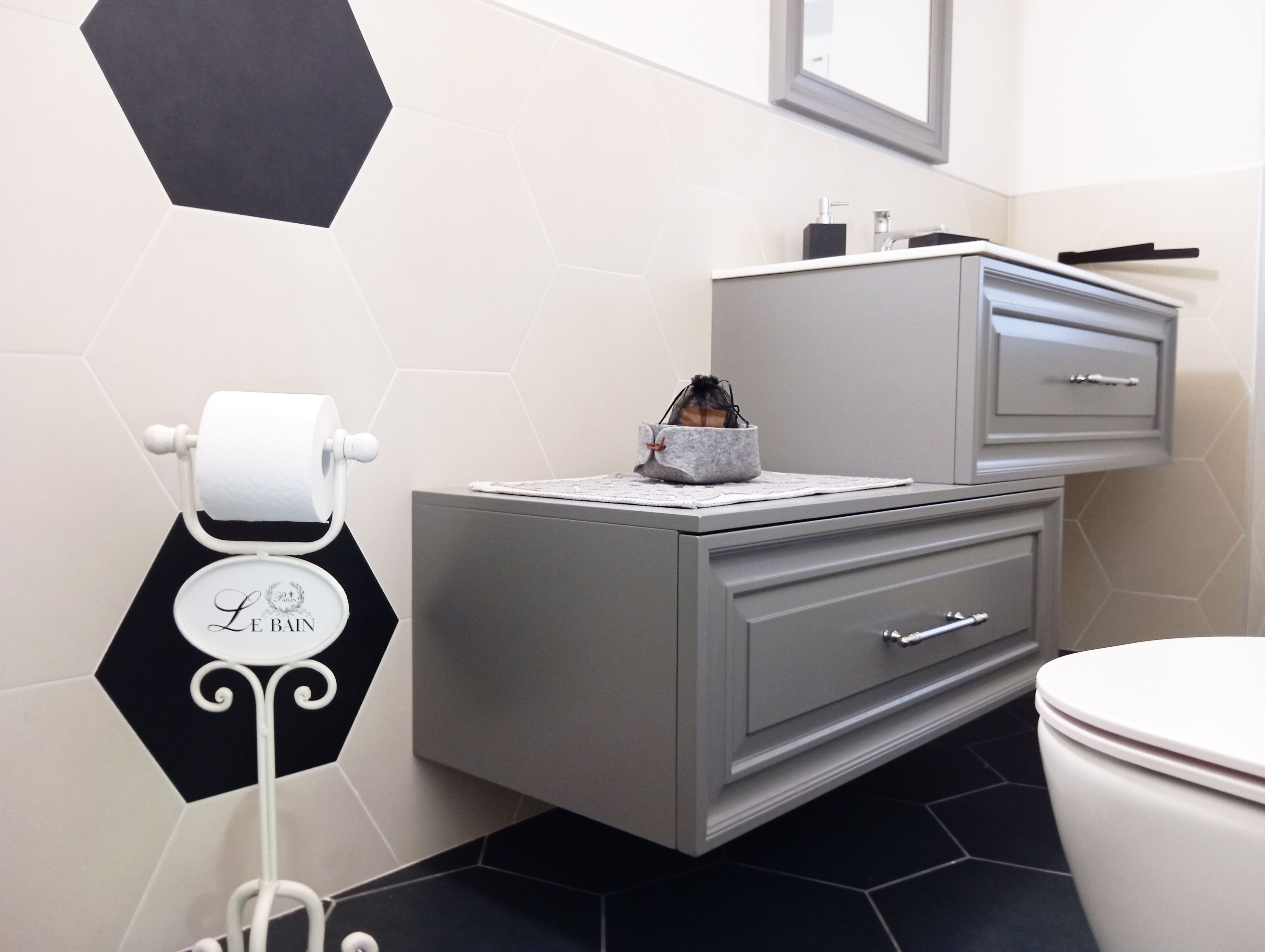 Une salle de bain comprenant des toilettes, un meuble-lavabo gris avec des tiroirs, un motif hexagonal noir et blanc sur le mur, et un porte-rouleau de papier toilette avec une étiquette.