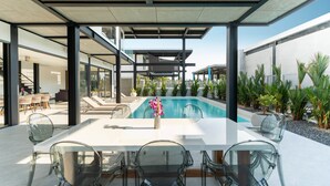 Un espace piscine extérieur moderne, doté d’une table et de chaises en verre, entouré de plantes et offrant une vue dégagée sur le ciel.
