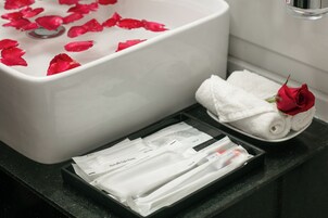 Un baño con lavamanos blanco, pétalos de rosa en el agua, una toalla enrollada con una rosa y artículos de aseo en una bandeja.