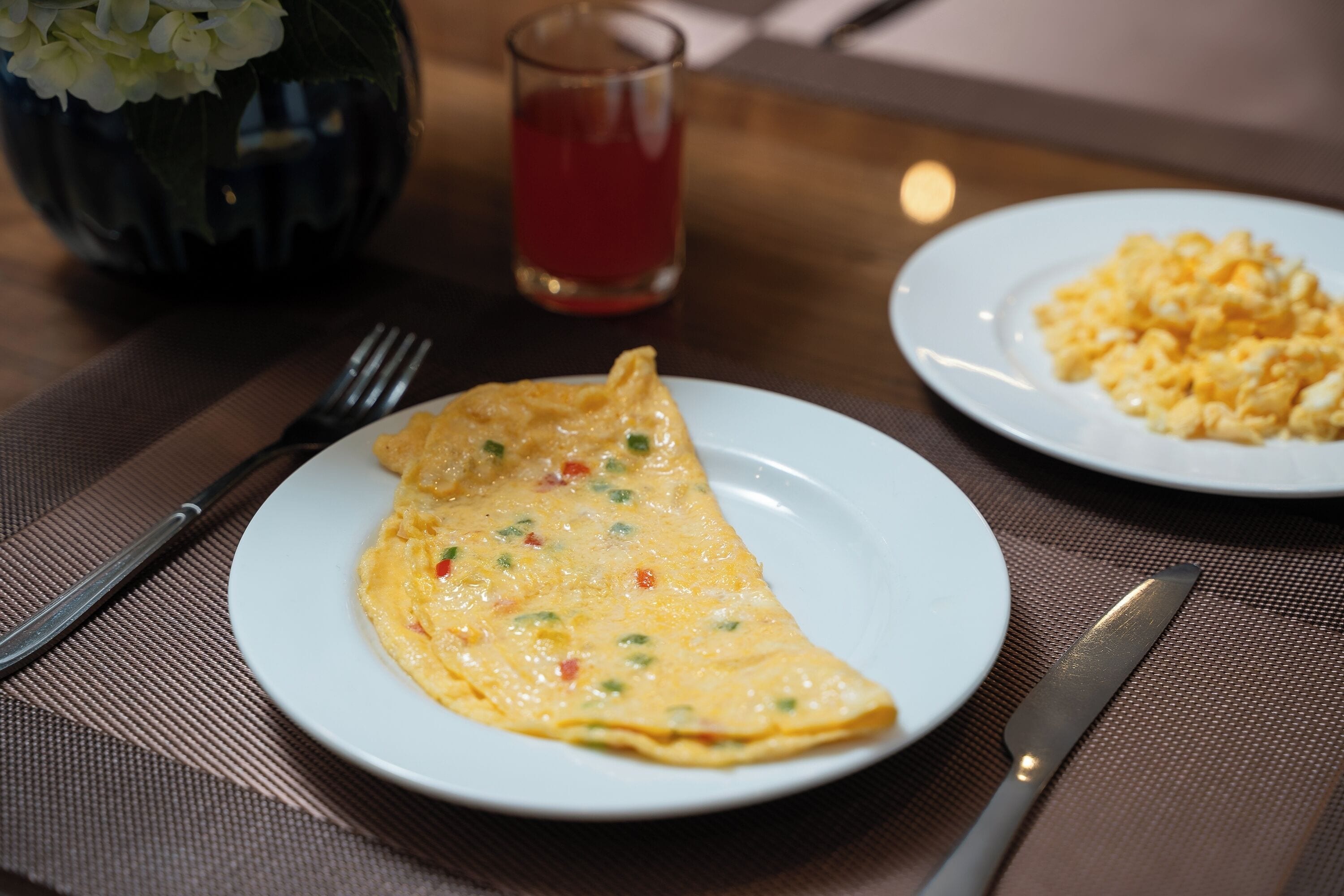 A plate of scrambled eggs and an omelette on a table with a glass of red drink and a vase of flowers in the background.