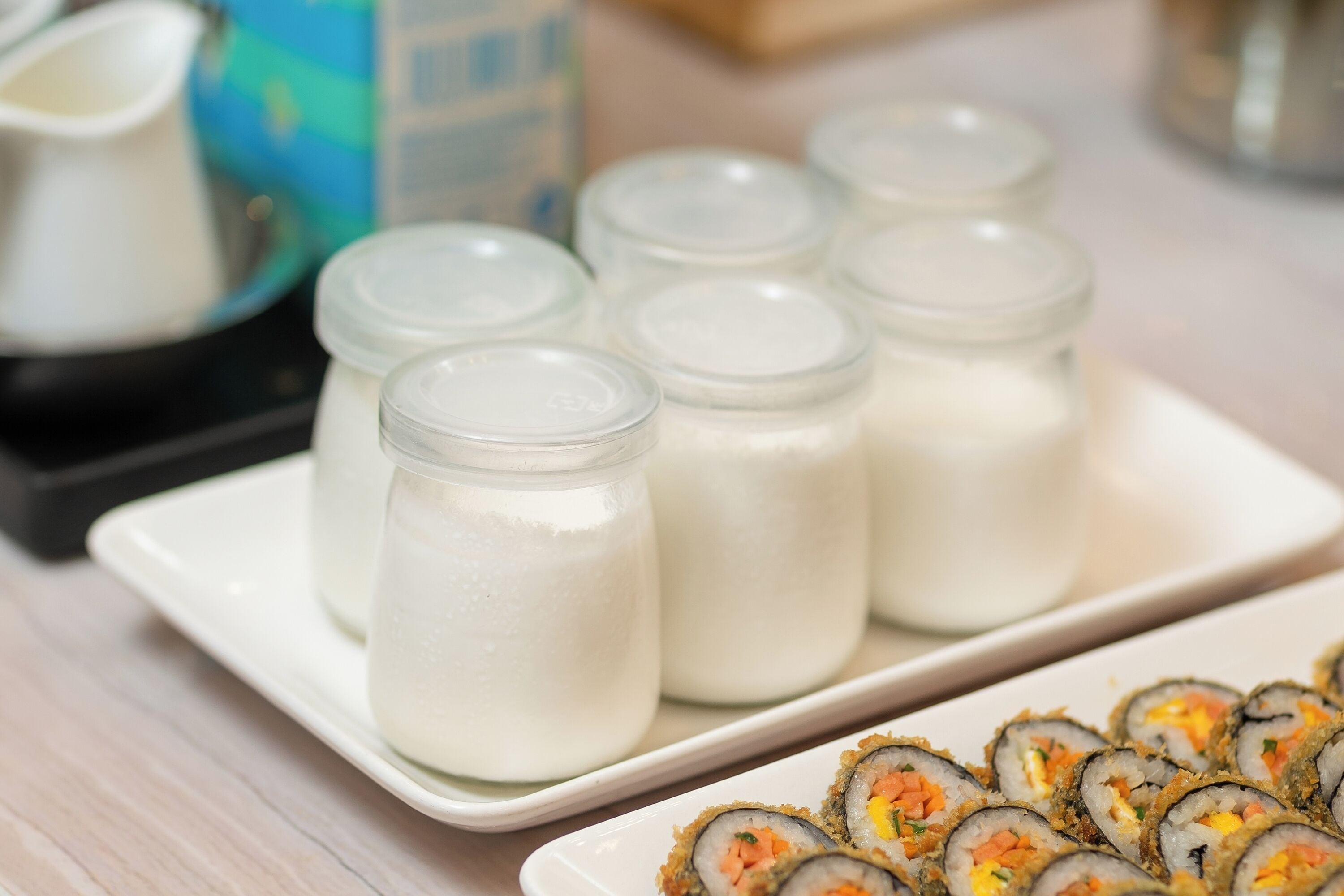 A plate of sushi and several glass jars of a white liquid, possibly milk or cream, on a white tray.