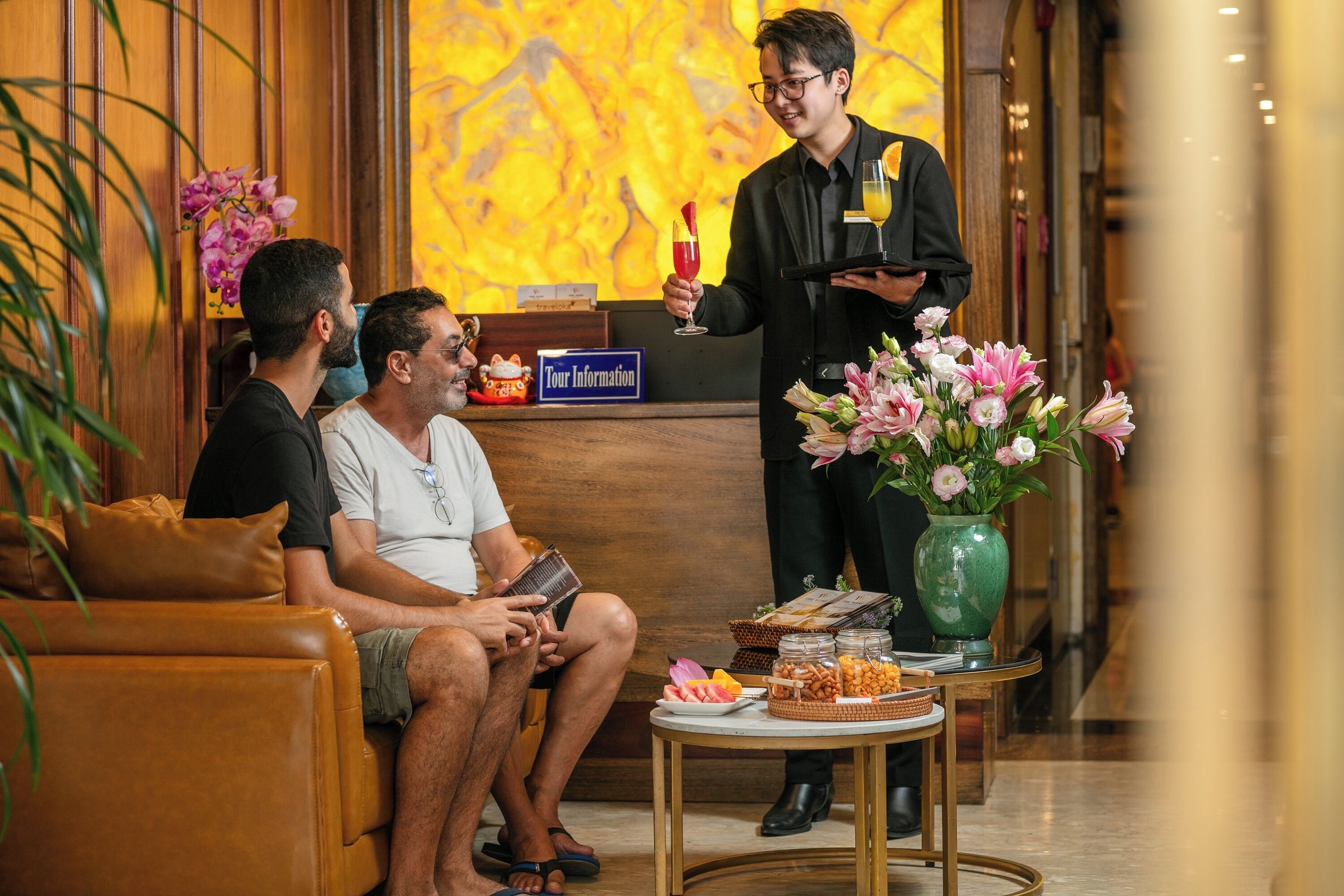 Zona de recepción de un hotel, con mostrador de recepción, un cartel que indica 'Tour Information' y una mesa con aperitivos y flores.