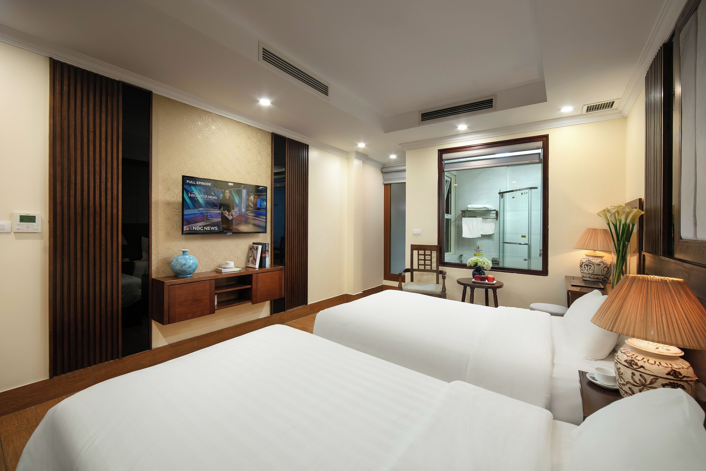A hotel room with two beds, a TV mounted on the wall, a mirror, a small table with a lamp, and a vase with flowers.
