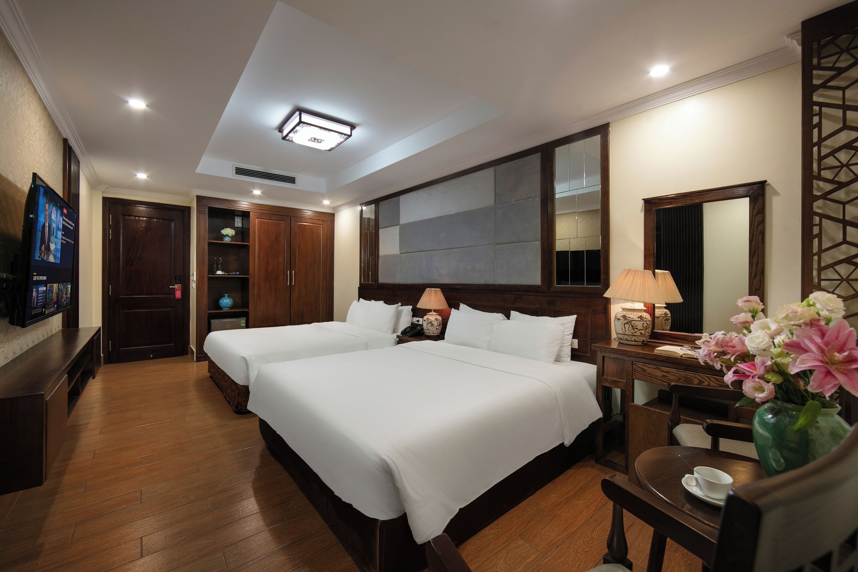 A hotel room with two beds, a television, a desk with a lamp, a vase of flowers, and a large mirror.