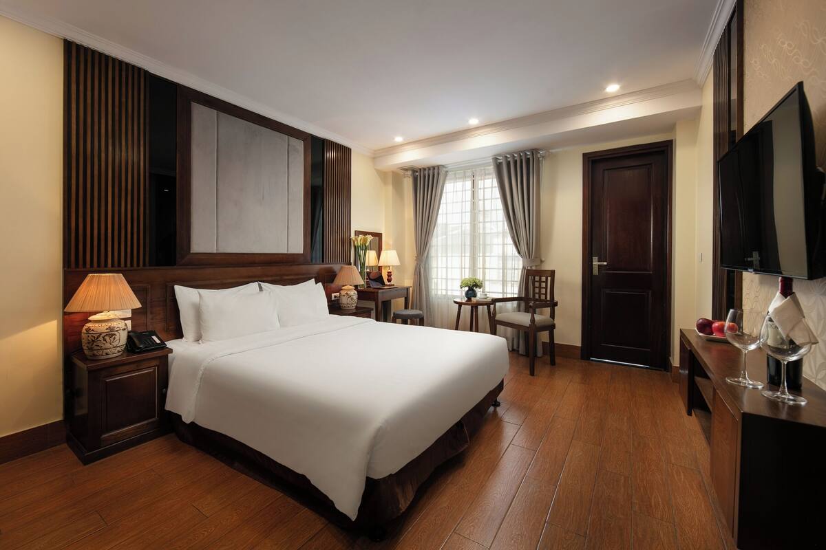 A hotel room with a large bed, bedside table with a lamp, a small table with a vase, a chair, a desk with a computer, and a TV mounted on the wall.