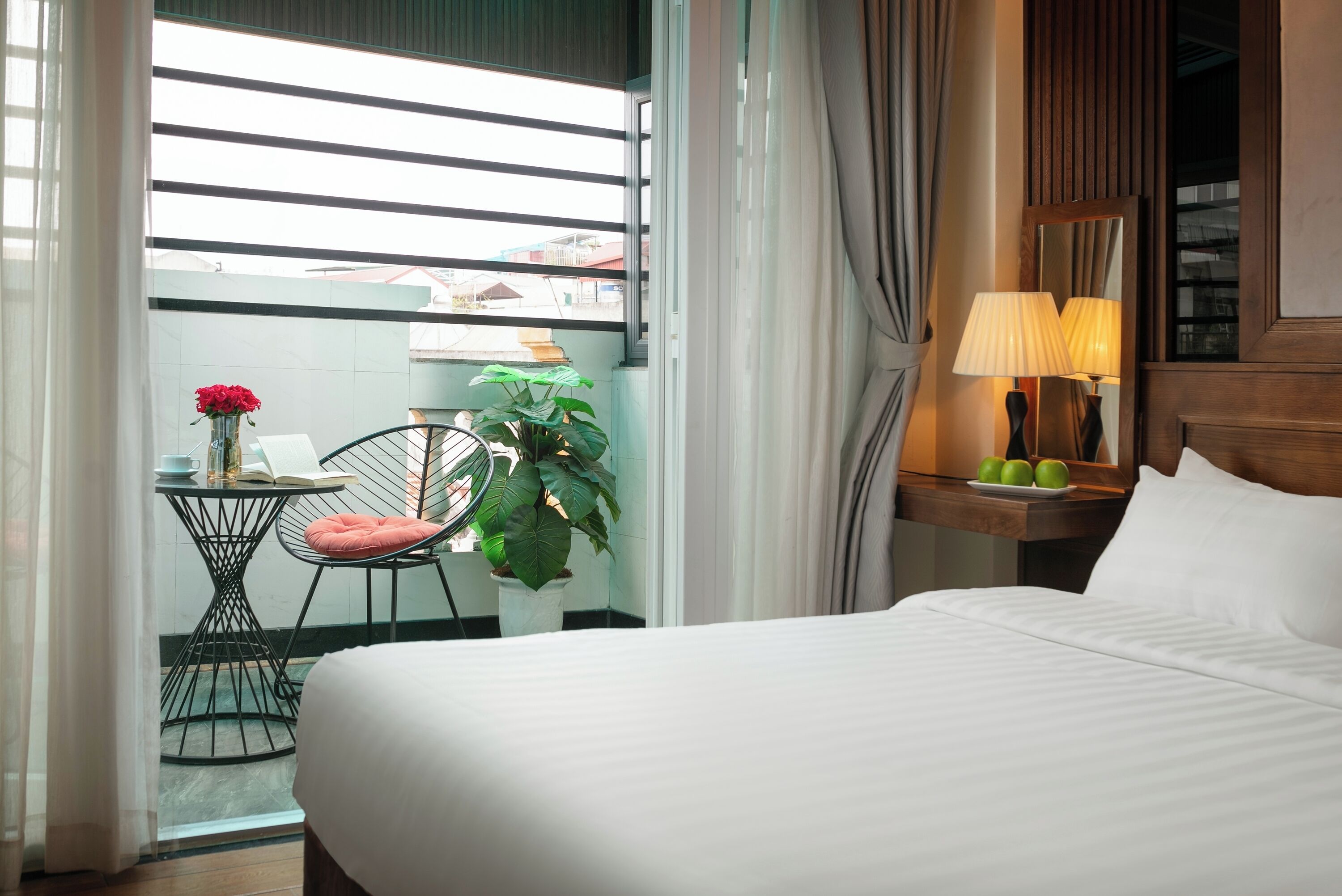 A hotel room with a bed, a small table with a chair, a potted plant, a lamp, and a window with curtains.