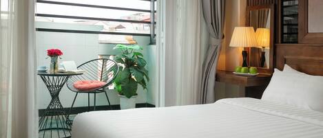 A hotel room with a bed, a small table with a chair, a potted plant, a lamp, and a window with curtains.
