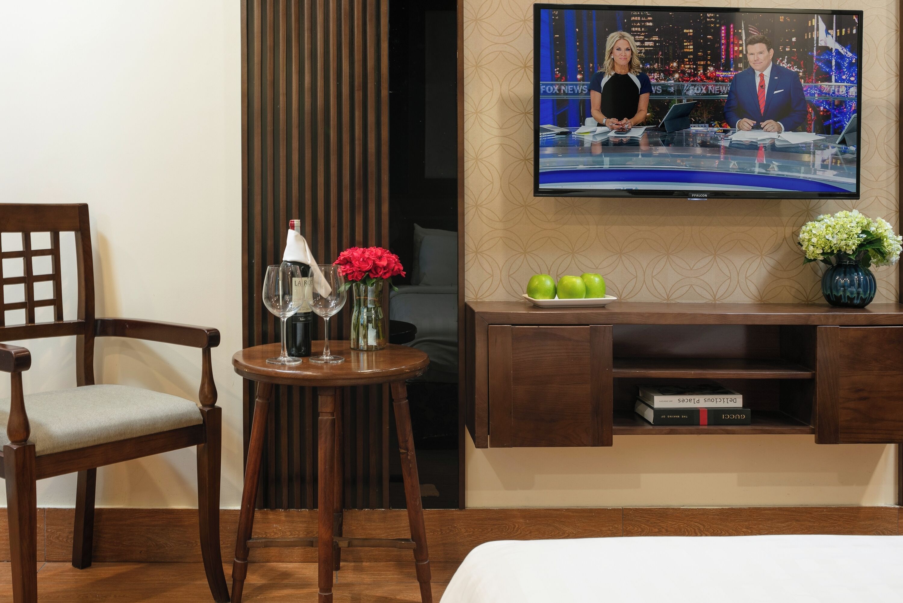 A hotel room with a wooden chair, a small round table with a vase of red flowers, a wine glass, and a flat-screen TV displaying a news broadcast.