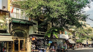 A street view with a building featuring a large tree, storefronts, and a clear sky. - The West Hotel & Spa (Hanoi)