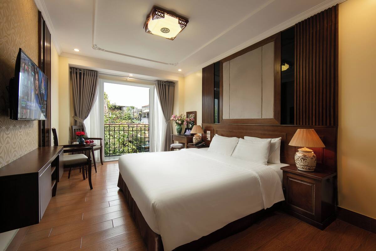 A hotel room with a large bed, a desk with a chair, a TV mounted on the wall, a nightstand with a lamp, and a view of the outdoors through a window.