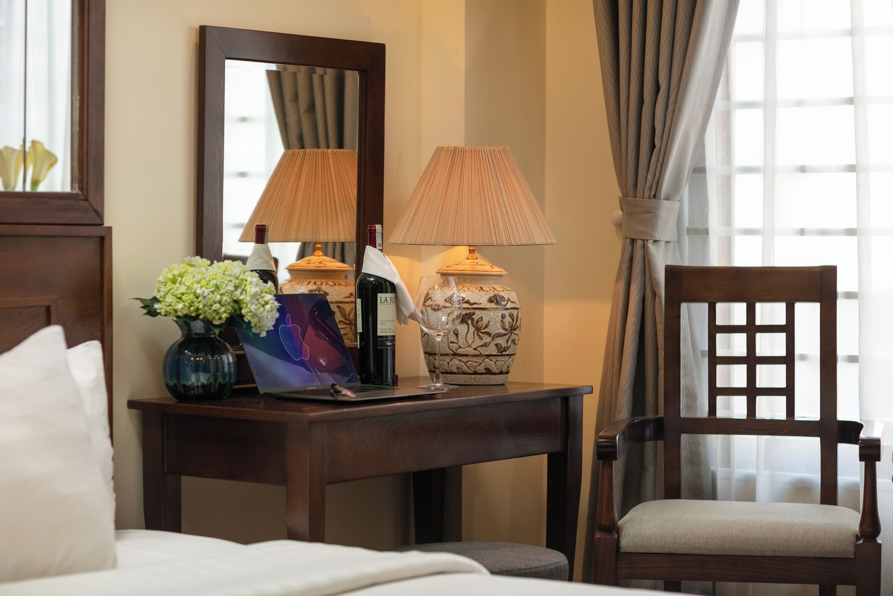 A hotel room with a bed, a wooden nightstand with a lamp, a vase with flowers, and a chair.