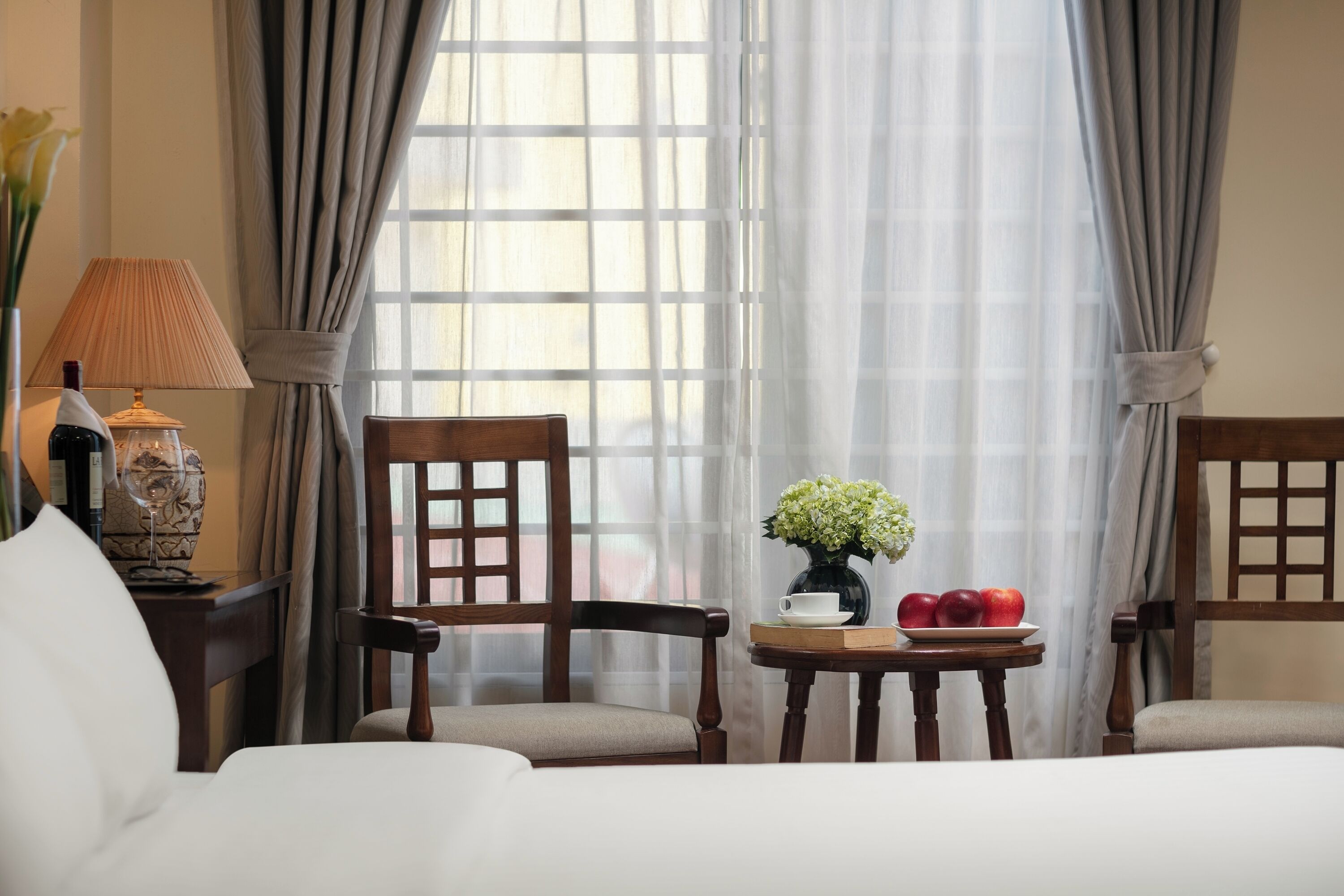 A hotel room with a bed, two wooden chairs, a small table with a vase of flowers and apples, and a lamp.