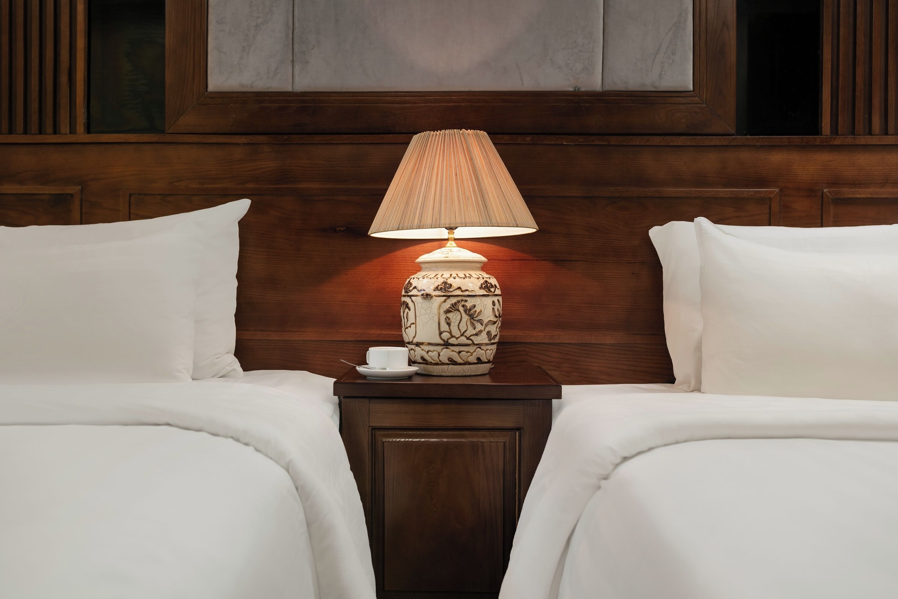 Quarto de hotel com cabeceira de madeira, duas camas brancas e uma mesa de cabeceira com abajur e uma chávena.