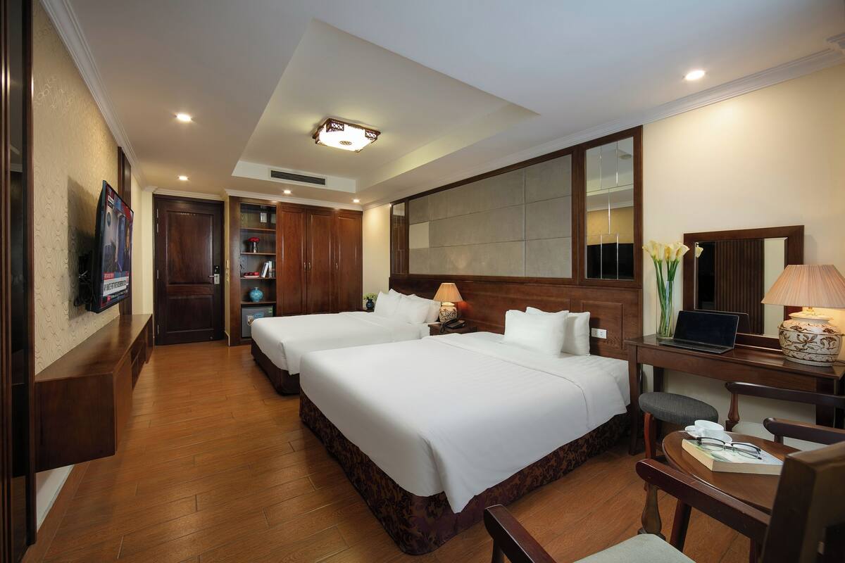 A hotel room with two beds, a desk, a chair, a lamp, a laptop, and a mirror.