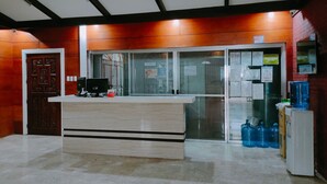 A reception area with a wooden reception desk, a computer, and a glass door with a metal gate. There are water dispensers and a refrigerator on the right. - Mang Ben Dormitory - Kaliraya QC - Hostel (Quezon City)