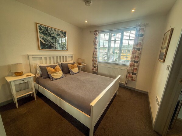 A bedroom with a bed, nightstands, a window with curtains, a radiator, and a painting on the wall. - The Crows Nest - Sheringham (Sheringham)