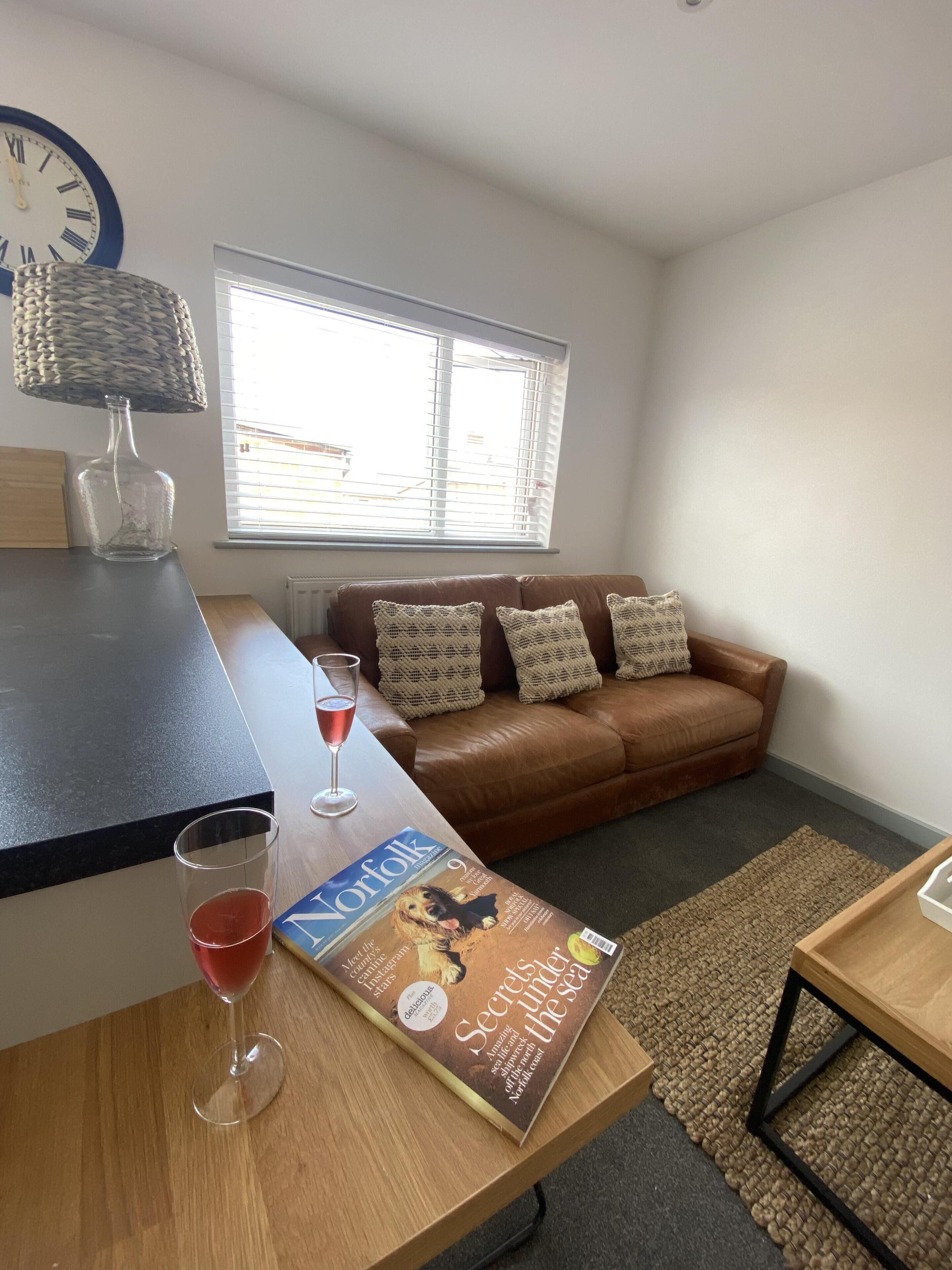 Ein Wohnzimmer mit einem braunen Ledersofa, einem Holztisch, einer Zeitschrift, einem Glas roter Flüssigkeit und einer Uhr an der Wand.