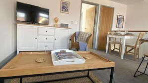 A modern living room with a flat-screen TV, a white cabinet, a dining table with chairs, and a woven rug. - The Crows Nest - Sheringham (Sheringham)