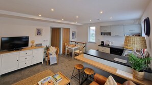A modern, well-lit living room with a TV, seating area, and a kitchen with a dining table. - The Crows Nest - Sheringham (Sheringham)
