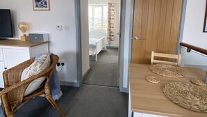 A room with a dining table, a wicker chair, a television, and a bed in the background. - The Crows Nest - Sheringham (Sheringham)