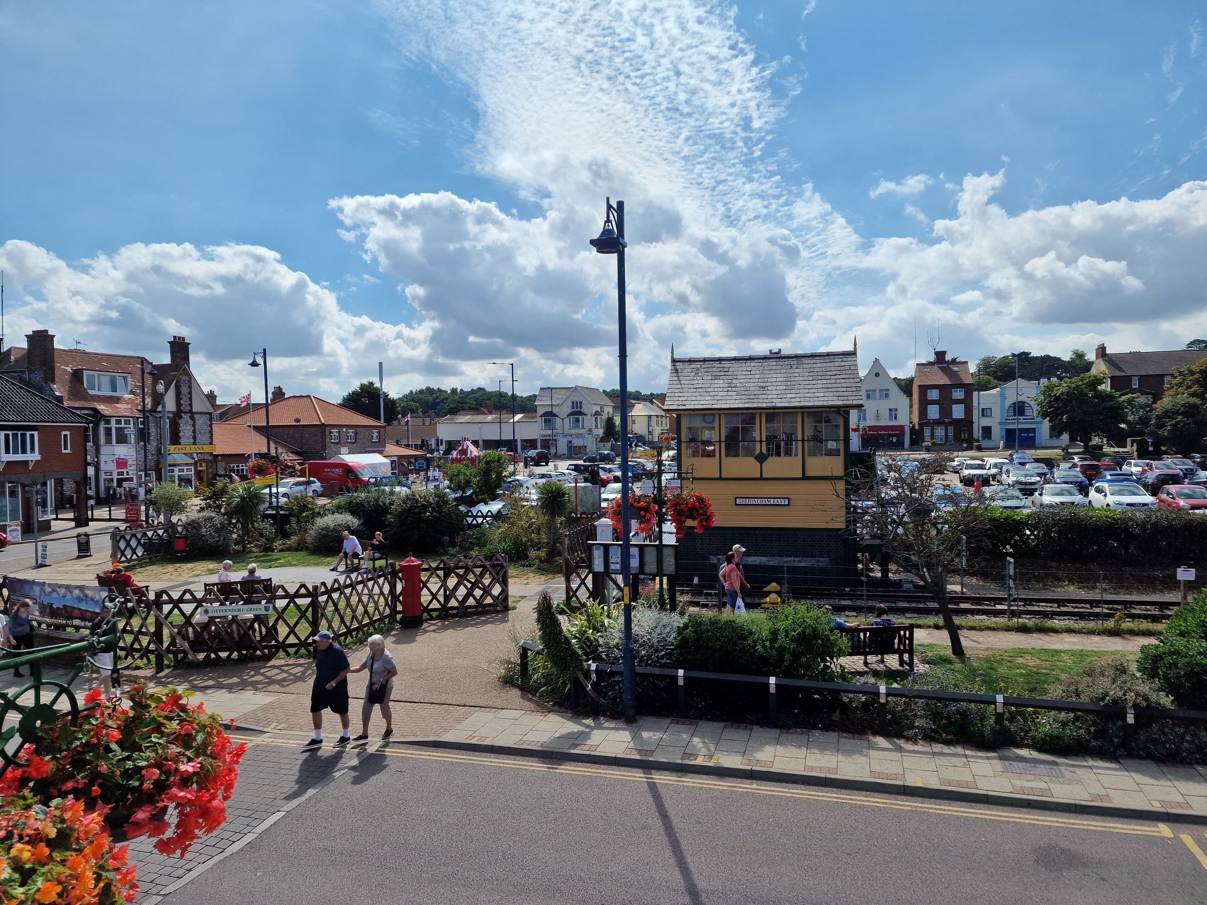 A town square with a railway signal box, parked cars, and people walking.