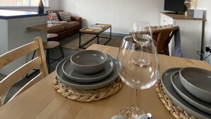 A dining area with a wooden table set for two, a framed crab artwork on the wall, and a kitchen area with a TV and a shelf. - The Crows Nest - Sheringham (Sheringham)