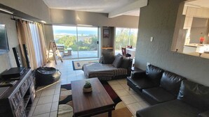 Uma sala de estar com um sofá, uma mesa de centro e uma televisão. Há uma varanda com vista para o oceano.