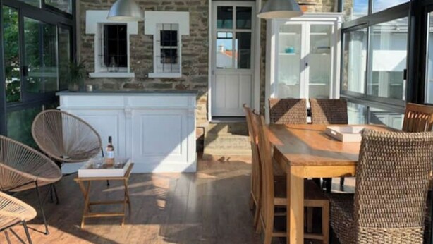 Een ruim terras met een houten tafel en stoelen, een stenen muur en grote ramen.