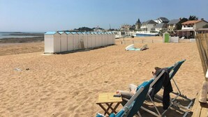 Een strand met een rij strandhutten, enkele mensen ontspannen op strandstoelen, en huizen op de achtergrond.