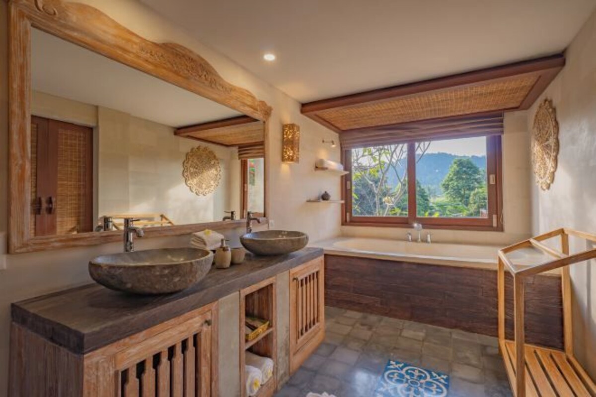 Un baño con un espejo grande, dos lavamanos, una bañera y una ventana con vistas a la vegetación.
