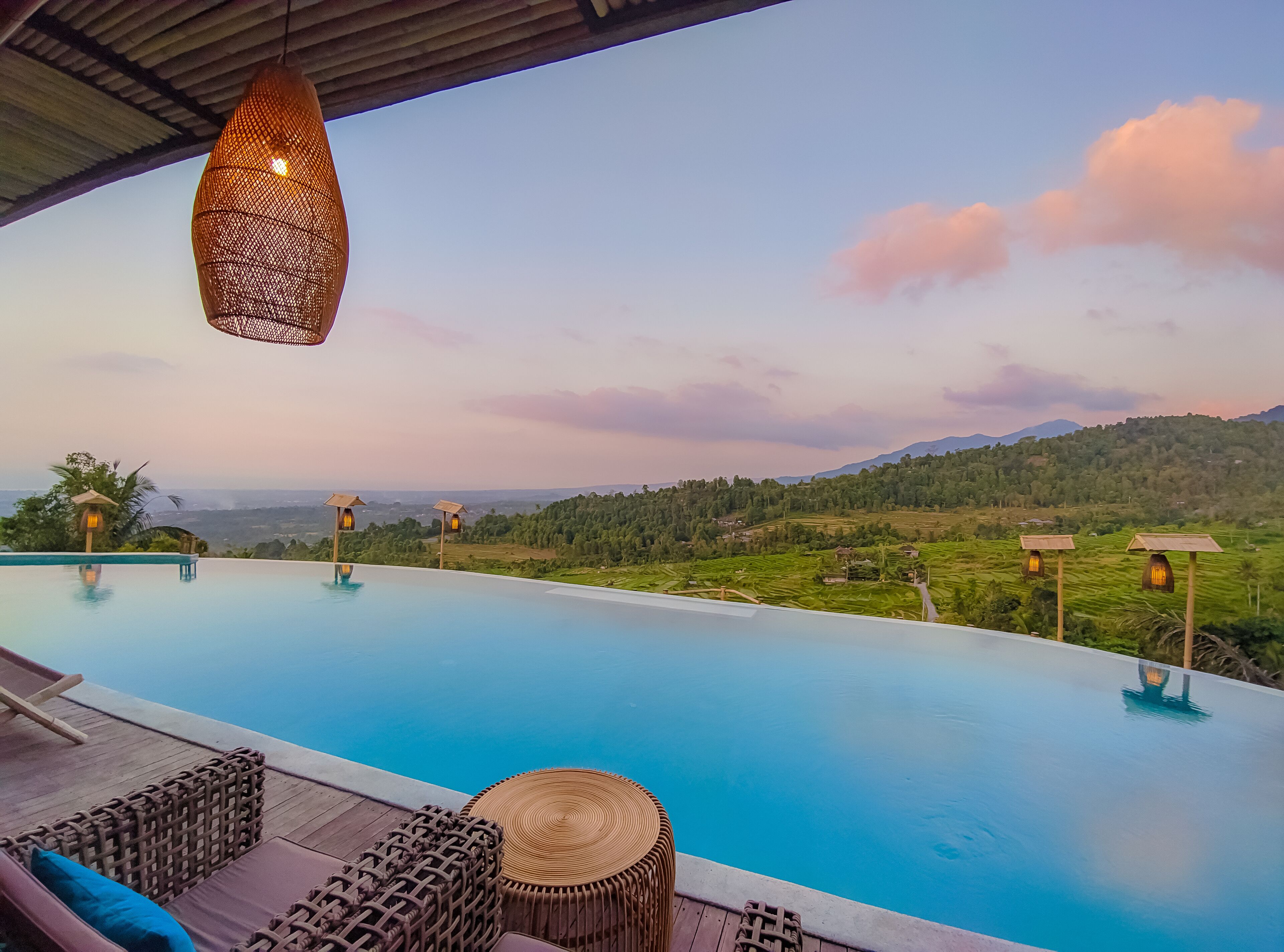 An infinity pool with wicker furniture and hanging lights, overlooking a lush valley and distant mountains.
