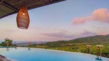 An infinity pool with wicker furniture and hanging lights, overlooking a lush valley and distant mountains.