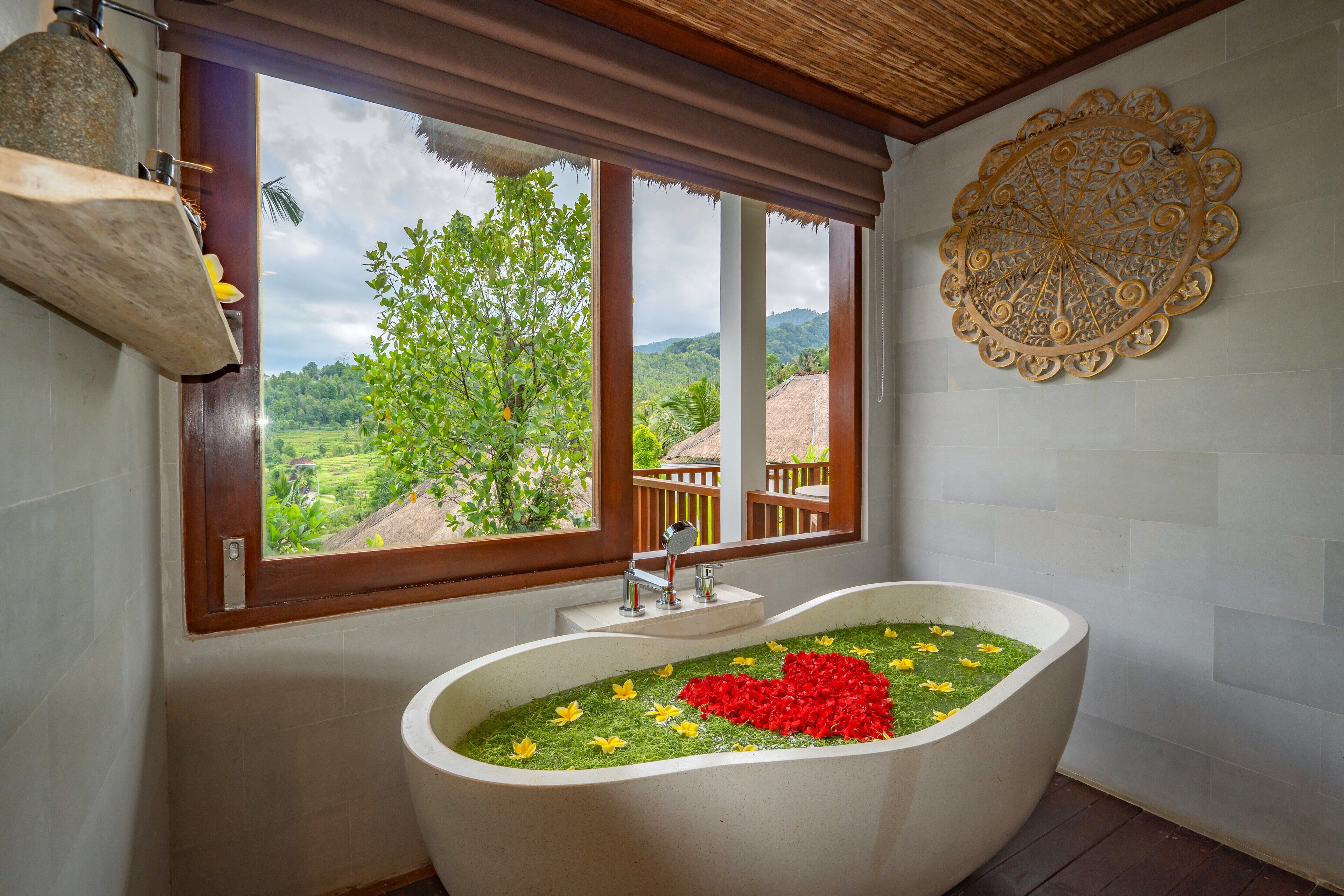 A bathroom with a bathtub filled with water and flowers, a window with a view of greenery, and a decorative wall piece.