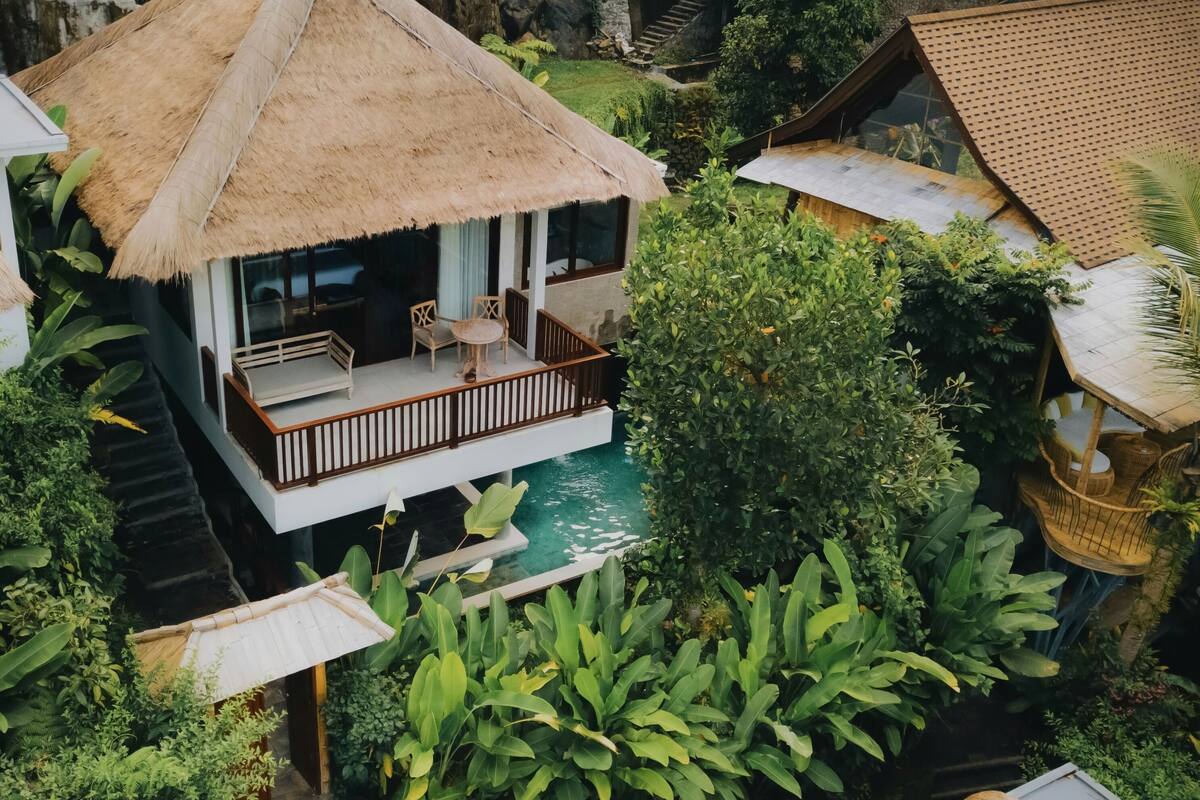 A bungalow with a thatched roof, a pool, and lush greenery.