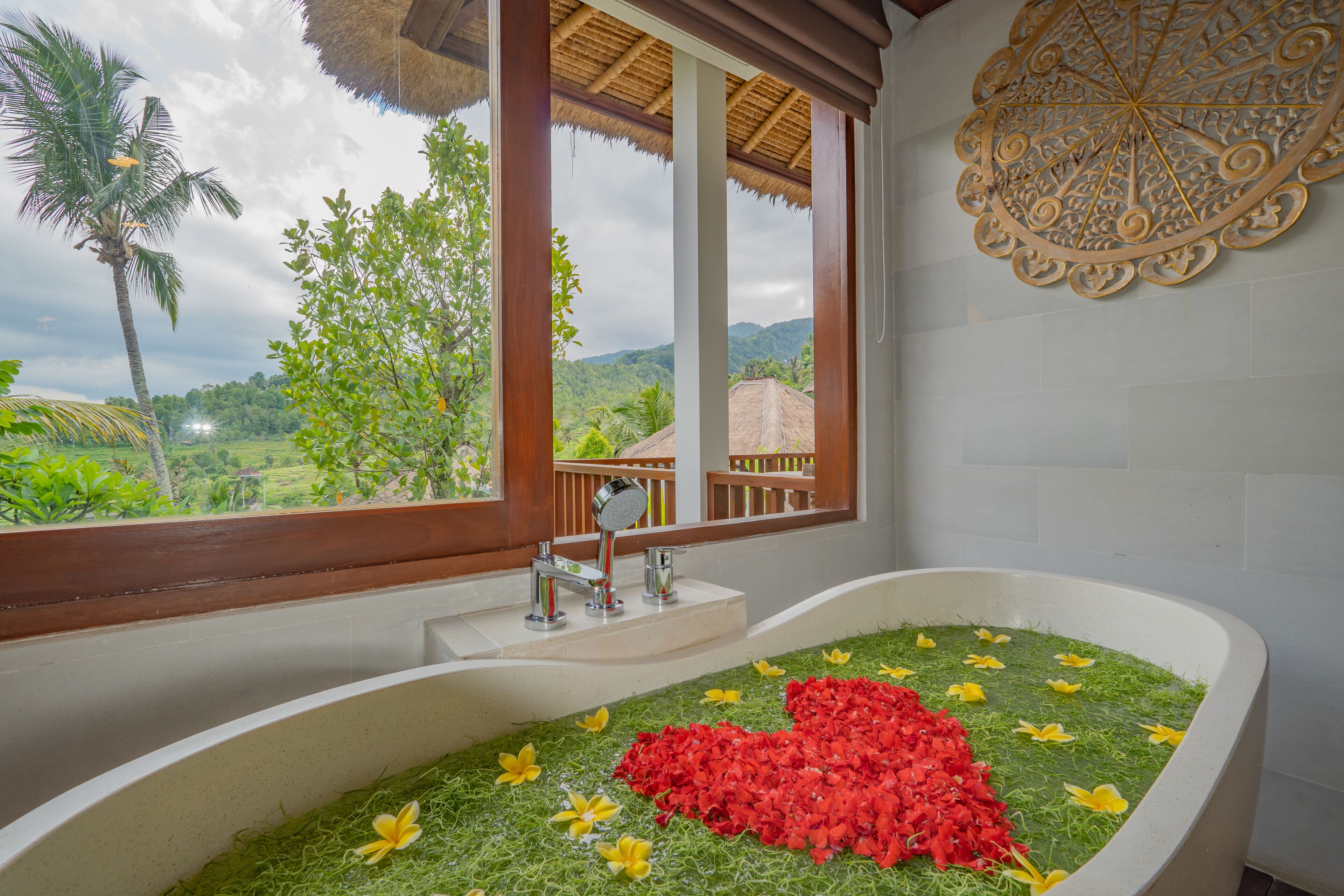 Un baño con una bañera grande llena de agua y flores, una ventana con vistas a la vegetación y un adorno en la pared.