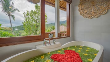 Un baño con una bañera grande llena de agua y flores, una ventana con vistas a la vegetación y un adorno en la pared.