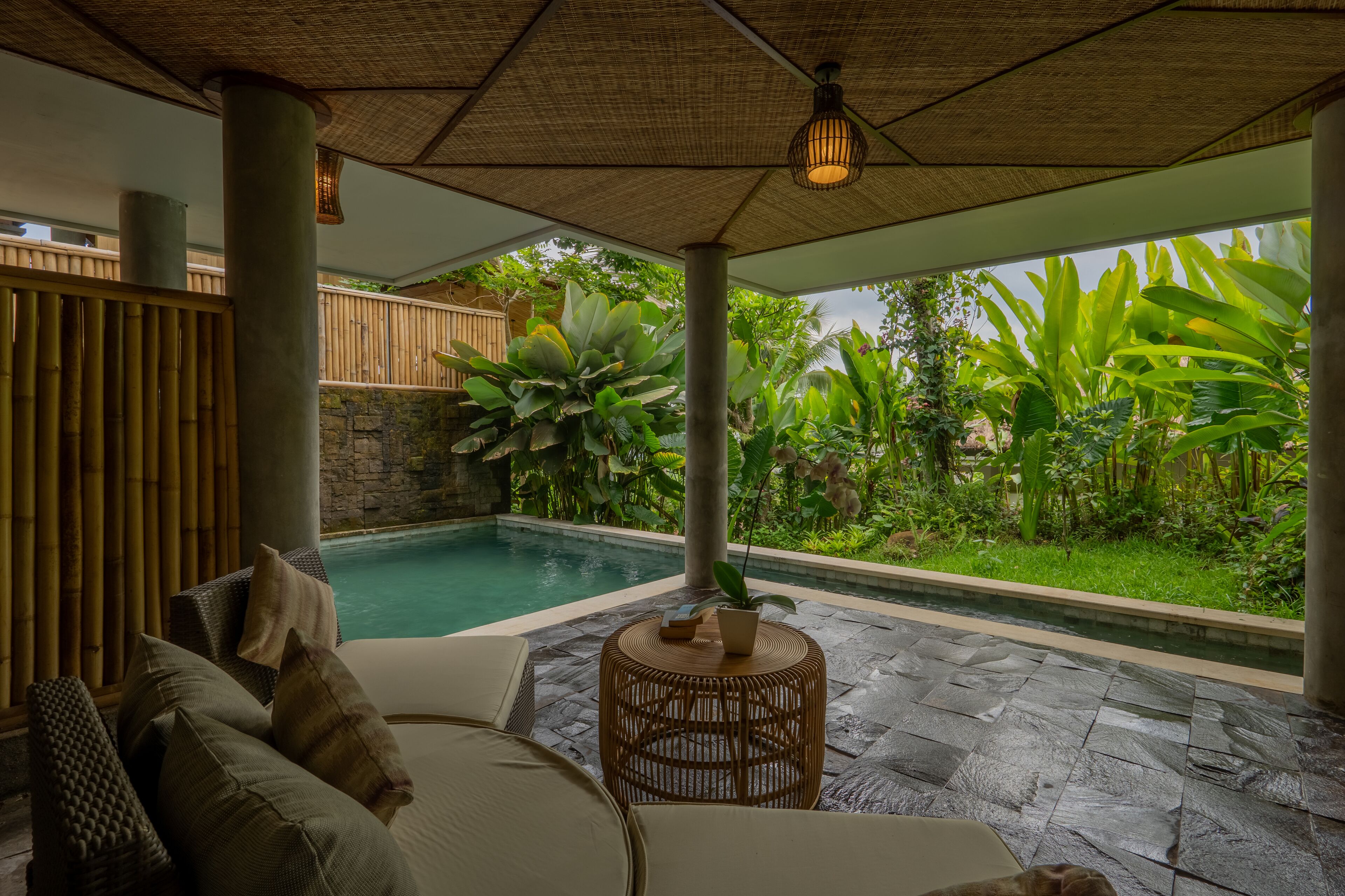 Un espace aménagé au bord de la piscine, avec un sol en pierre, une chaise en osier et une table agrémentée d’une plante.