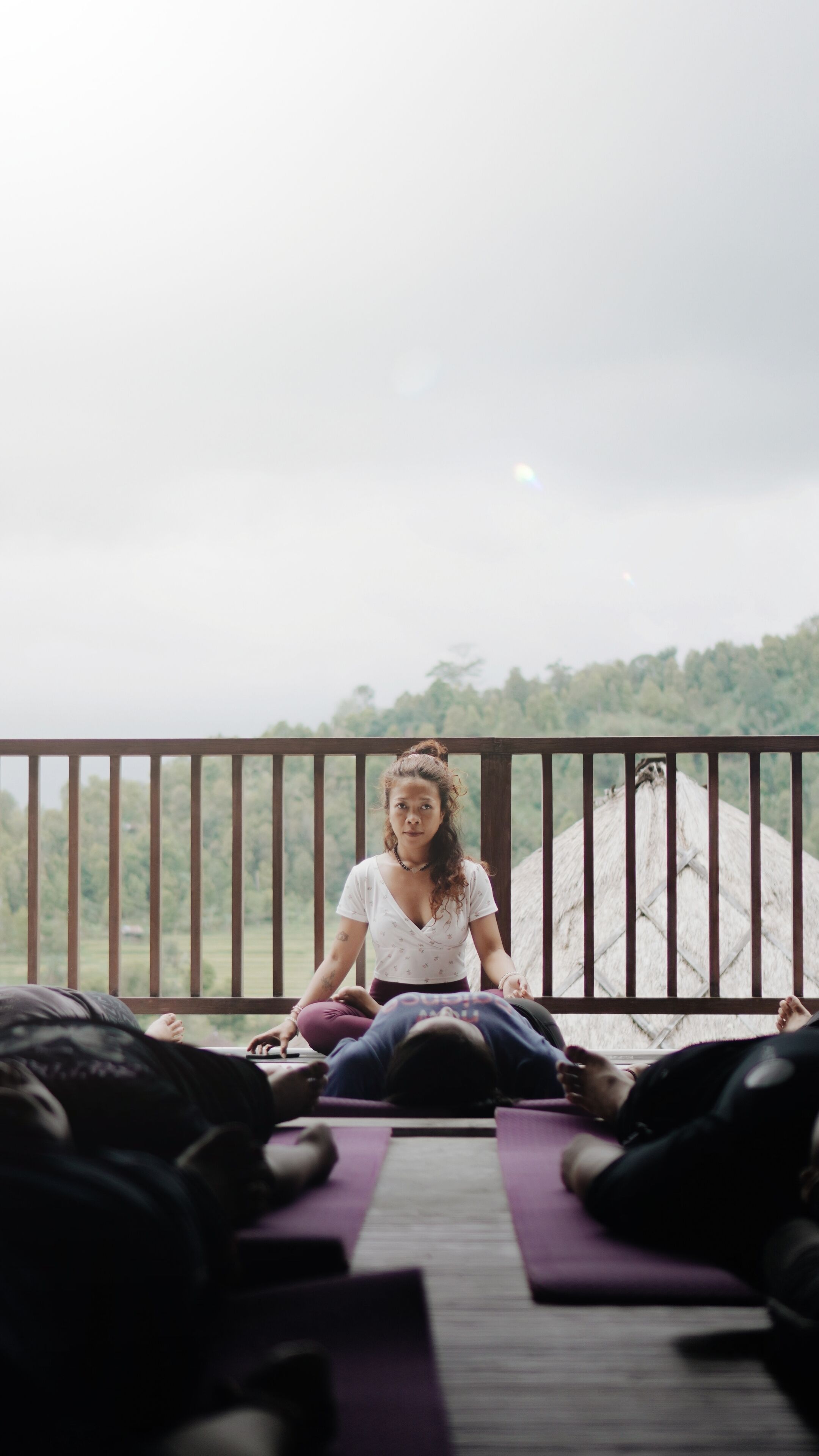 Une personne assise sur un tapis de yoga à l’extérieur, avec une rambarde derrière elle et une colline boisée en arrière-plan.