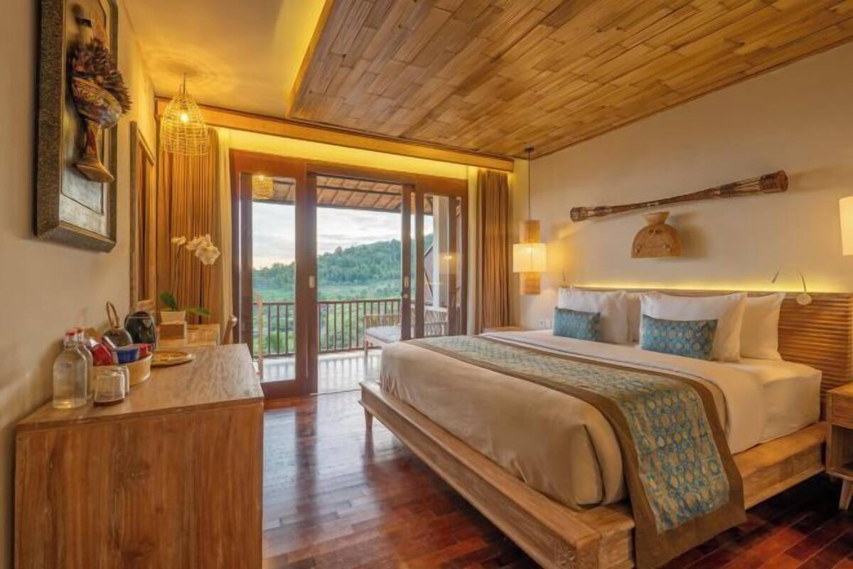 A bedroom with a large bed, wooden headboard, bedside lamps, a wooden dresser, and a view of the outdoors.
