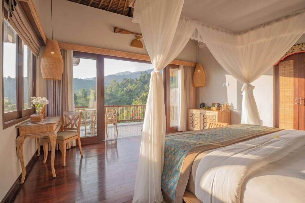 A bedroom with a large bed, a balcony with a view, and a wooden desk with chairs.