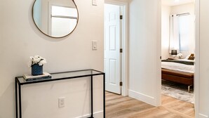 A hallway with a round mirror, a glass console table with a vase of flowers, and a white door leading to a bedroom with a bed and a window. - LOVELY STAY |GREAT LOCATION NEAR DISNEY & DOWNTOWN (Fullerton)