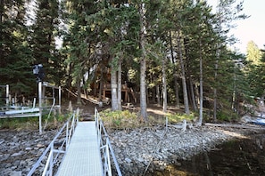 Una cabaña de madera rodeada de altos árboles, accesible a través de un puente metálico sobre un arroyo pedregoso.