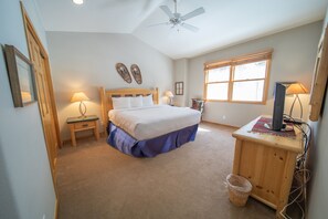 Quarto com cama, luminárias de cabeceira, ventilador de teto, janela e escrivaninha com computador.